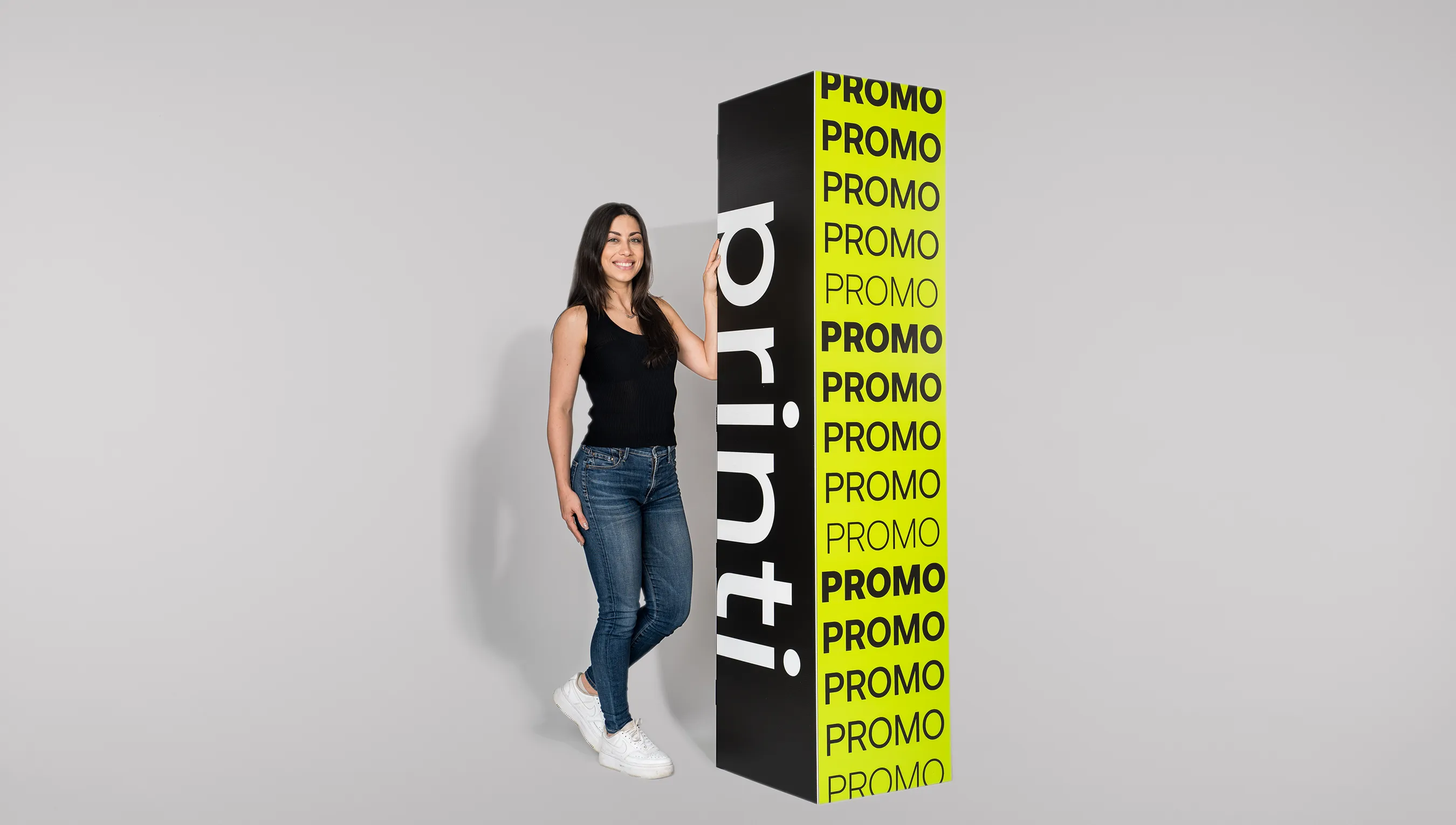 otem quadriedro da Printi em preto e amarelo com uma mulher branca usando calça jeans, camiseta preta e tênis branco apoiada em pé ao lado do totem