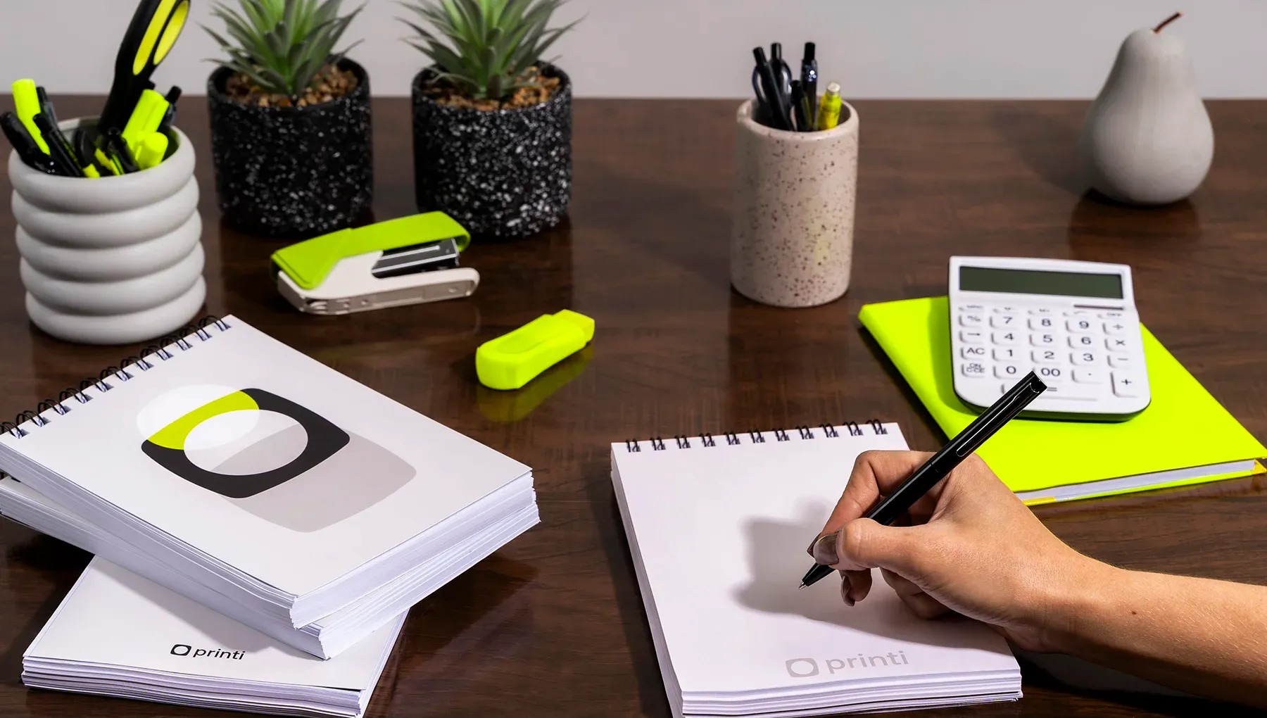 Mesa de escritório organizada com mão escrevendo em um bloco de notas com capa wire-o branco da Printi, cercado por calculadora, grampeador verde neon e acessórios de papelaria.