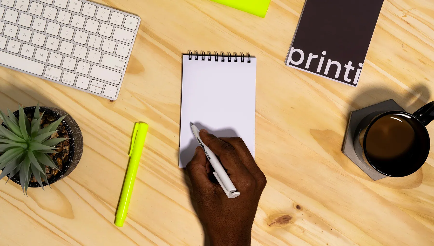 Pessoa escrevendo em um bloco para rascunho espiral branco sobre mesa de madeira com teclado, café e planta ao lado.