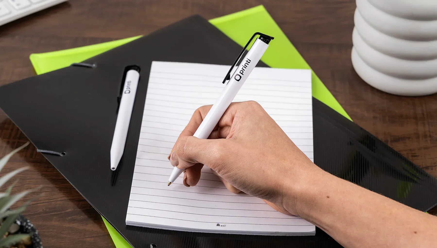 Mão escrevendo em bloco de notas com caneta plástica básica personalizada na cor branca e detalhes pretos, ideal para brinde corporativo. Ao fundo, pasta de escritório.