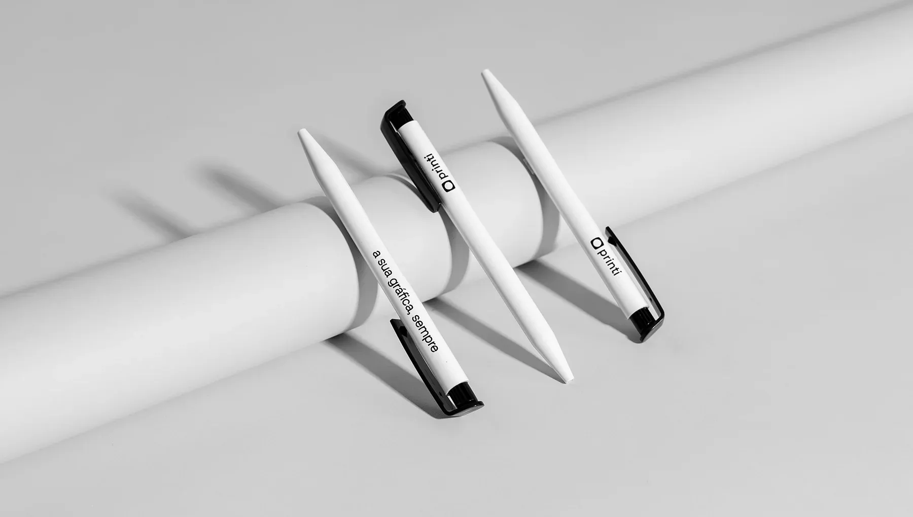 Três unidades de caneta plástica básica personalizada com logotipo e a frase institucional "a sua gráfica, sempre", exibidas em cenário minimalista.